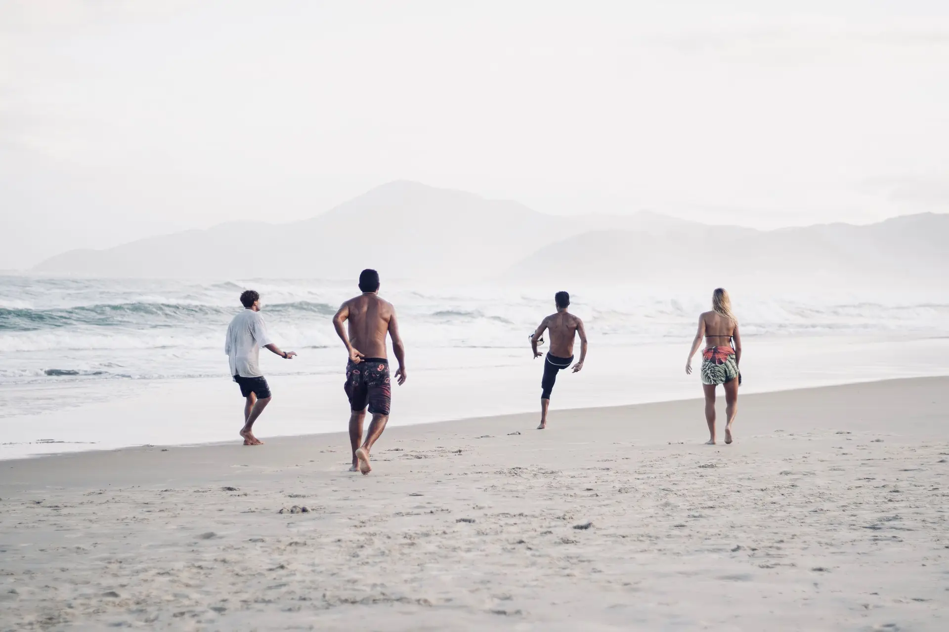A group of adults playing soccer on a sandy beach with mountains in the background.