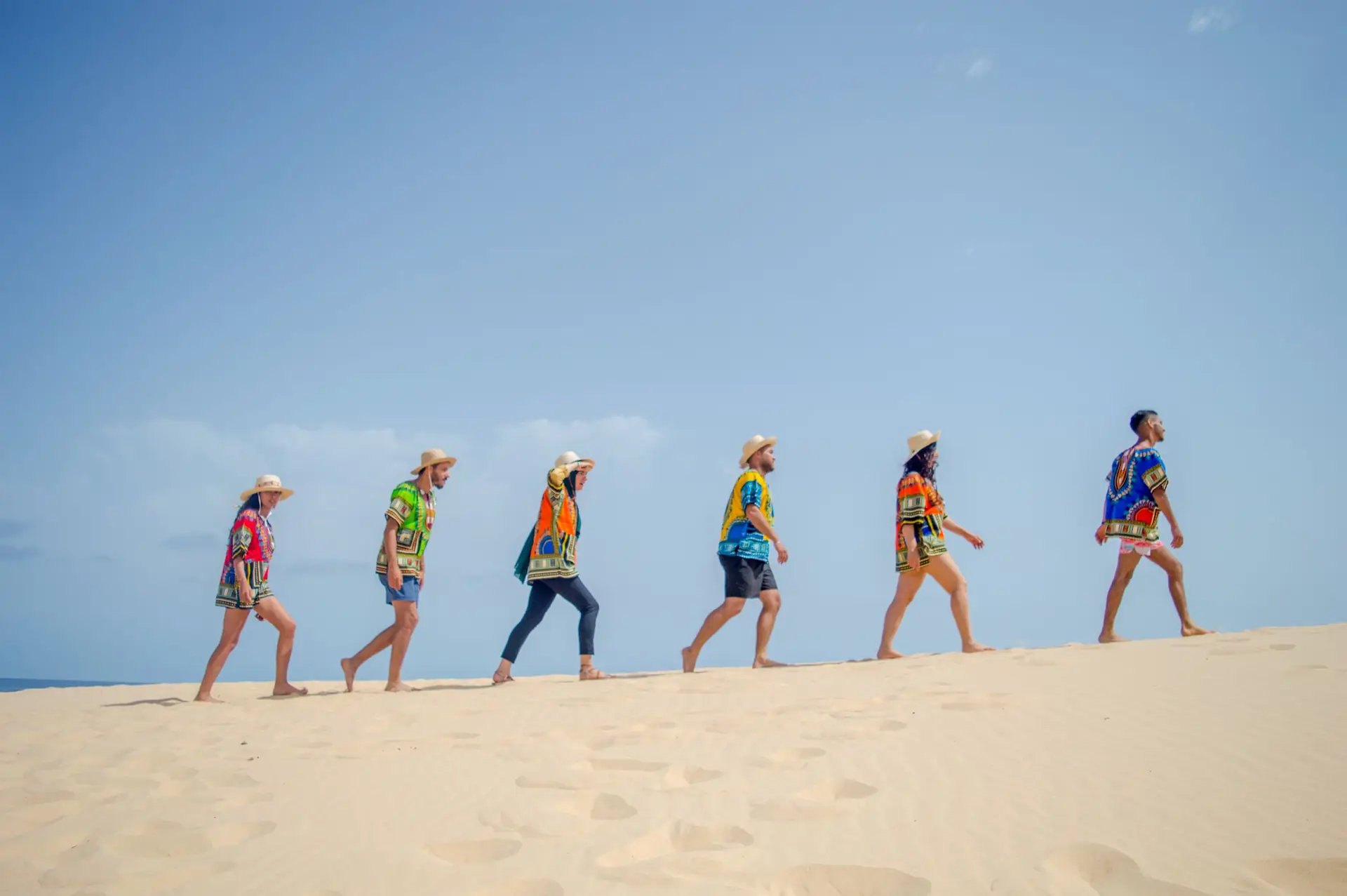 Side view group of anonymous young diverse travelers in colorful summer outfits walking on sandy beach near sea against blue sky